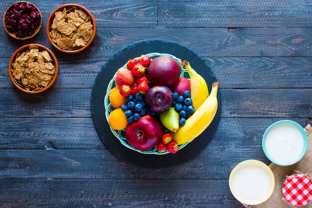 Bol de fruits frais avec banane, pomme, fraises, abricots, myrtilles, prunes, grains entiers, fourchettes, vue de dessus