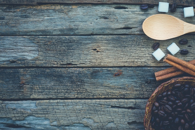 Bol de grains de café torréfiés et cuillère avec des bâtons de cannelle et des morceaux de sucre sur la table en bois