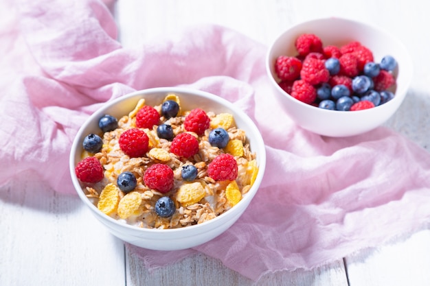 Photo bol avec granola, bleuets et framboises dans un bol blanc, tissu rose sur une surface blanche. concept de petit déjeuner sain