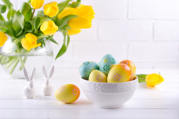 Bol avec des oeufs de Pâques colorés, décoration de Pâques au printemps sur une table en bois blanc avec bouquet de fleurs de tulipes jaunes dans un vase en verre sur fond blanc. Décoration intérieure de Pâques