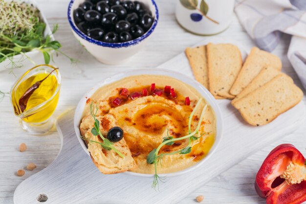 Un bol de pois chiches houmous maison avec de l'huile d'olive, du pain croustillant, microgreen et paprika fumé sur une surface en bois blanche.