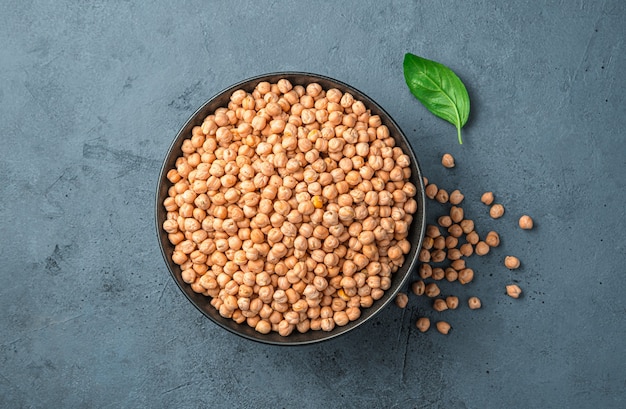 Un bol avec des pois chiches secs et crus sur un fond gris-bleu. Vue de dessus, gros plan.