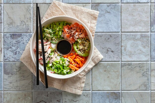 Bol Poke fait maison à la mode pan-asiatique avec saumon, riz, edamame, légumes frais et sauce soja, servi avec des baguettes sur fond de carreaux de céramique.