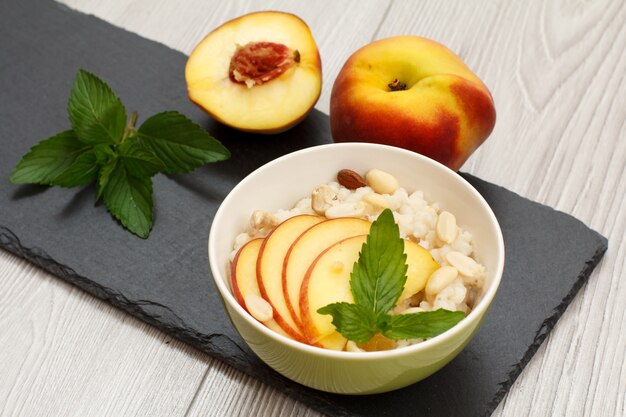 Bol en porcelaine avec bouillie de sorgho, pêche fraîche, noix de cajou, feuilles d'amande et de menthe sur planche de pierre et table en bois gris. Salade de sorgho vegan sans gluten aux fruits frais. Vue de dessus.