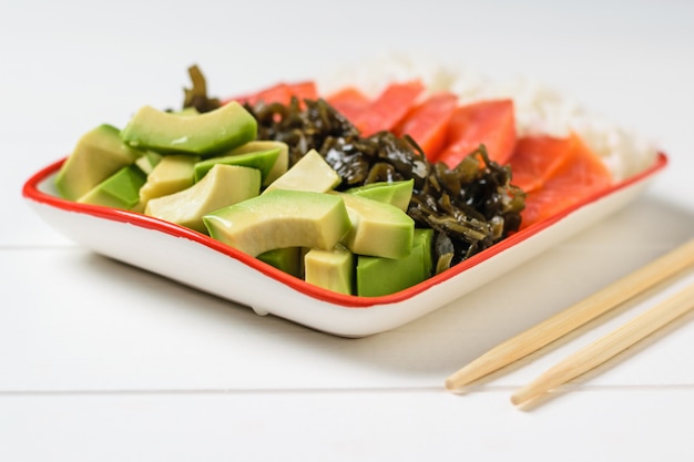 Bol avec riz, algues, tranches d'avocat et poisson sur une table blanche avec des bâtons en bois.