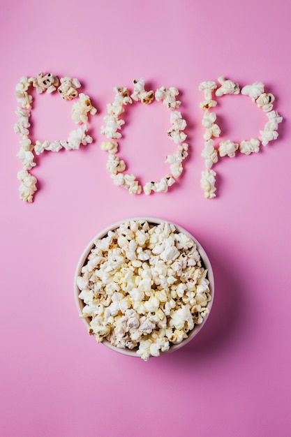 Un bol rose de pop-corn sur une surface rose avec mot POP en maïs