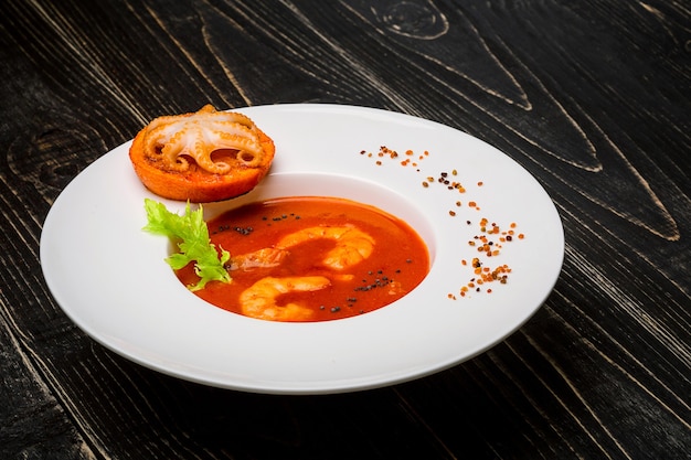 Bol de soupe de tomates aux crevettes avec une petite pieuvre cuite sur une tranche d'orange sur un bac en bois noir...