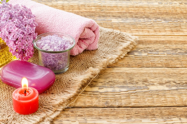 Bol en verre avec sel de mer, savon, bougie allumée, fleurs lilas et serviette pour les procédures de salle de bain sur des planches en bois. Produits et accessoires de spa. Vue de dessus.