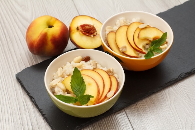 Bols en porcelaine avec bouillie de sorgho, pêches fraîches, noix de cajou, feuilles d'amande et de menthe sur une planche en pierre noire. Salade de sorgho vegan sans gluten aux fruits frais. Vue de dessus.