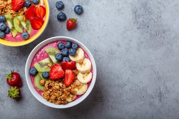 Bols de smoothie açai d'été avec fraises, banane, myrtilles, kiwis et granola sur fond de béton gris. Bol de petit-déjeuner avec fruits et céréales, gros plan, vue de dessus, espace pour le texte