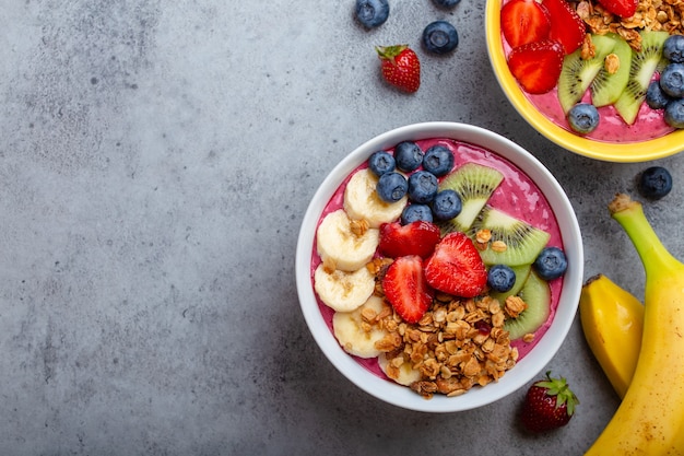 Bols de smoothie açai d'été avec fraises, banane, myrtilles, kiwis et granola sur fond de béton gris. Bol de petit-déjeuner avec fruits et céréales, gros plan, vue de dessus, espace pour le texte