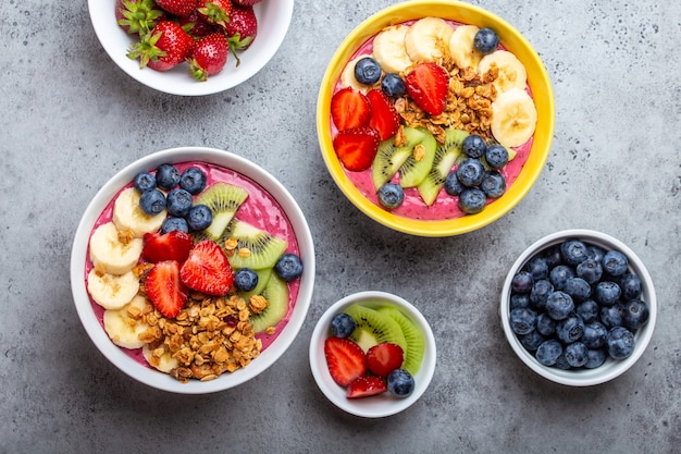 Bols de smoothie açai d'été avec fraises, banane, myrtilles, kiwis et granola sur fond de béton gris. Bol de petit-déjeuner avec fruits et céréales, gros plan, vue de dessus, nourriture saine