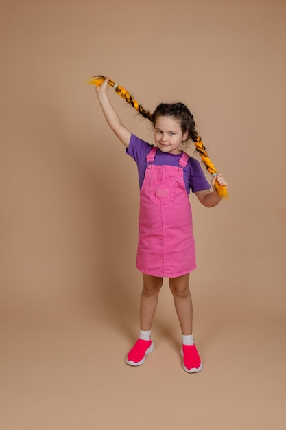 Bon jeune enfant obéissant jouant avec des tresses kanekalon de couleur jaune regardant la caméra légèrement souriant portant une combinaison rose et un t-shirt violet sur fond beige