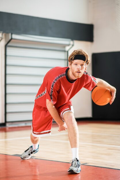 Bon joueur. Homme au gingembre dans un vêtement de sport rouge jouant au basket