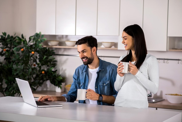 Bon moment Joyeux jeune couple buvant du thé et utilisant un ordinateur portable dans la cuisine