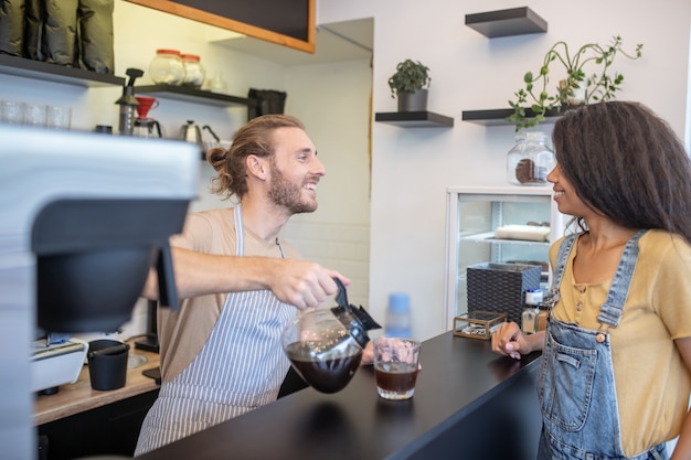 Bon service. Profil de jeune homme adulte affable verser le café de la cafetière derrière le comptoir et femme mulâtre en attente