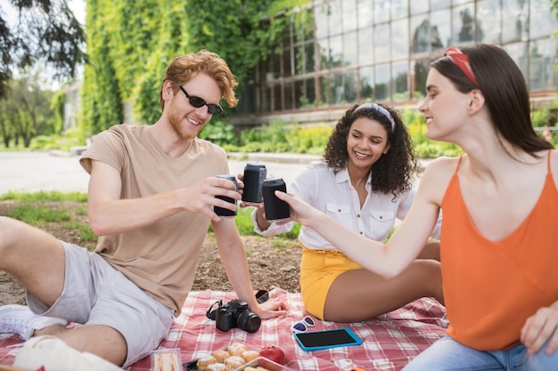 Bon temps. Groupe de jeunes gens optimistes et sympathiques se reposant assis sous un arbre sur une couverture avec un verre par une chaude journée dans le parc