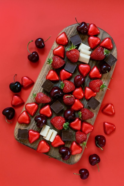 Bonbons en forme de coeur, chocolat noir et blanc, fraises et baies de cerises sucrées sur planche de charcuterie sur fond rouge. Gros plan, vue de dessus.