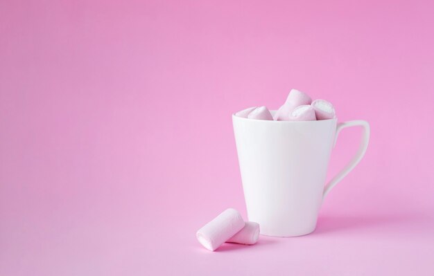 bonbons de guimauve rose dans une tasse blanche sur fond rose.