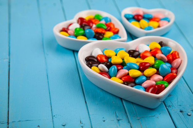 Bonbons multicolores dans une assiette en forme de coeur sur fond bleu en bois. La Saint-Valentin. Place pour votre texte.