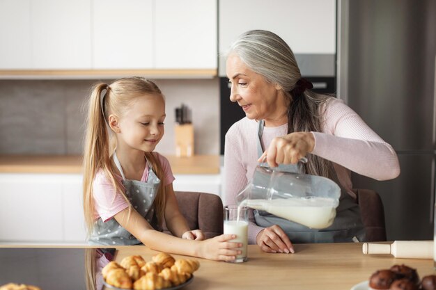 Les bonbons sont prêts Une grand-mère âgée européenne heureuse en tablier verse du lait au verre de sa petite-fille