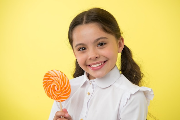 Bonbons sucette friandises préférées de l'élève S'amuser avec des bonbons Fille kid queues de cheval coiffure manger sucette sucrée Fille élève uniforme scolaire comme sucette sucrée fond jaune Sa saveur préférée