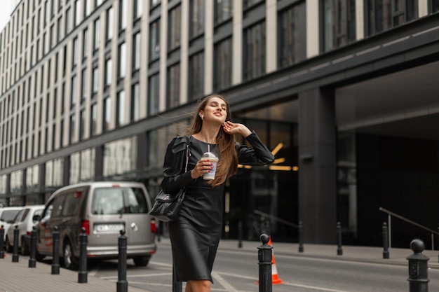 Bonheur élégante belle femme d'affaires en tenue noire de mode avec robe et sac en cuir tient une tasse de café et se promène dans la ville près d'un bâtiment moderne Fille souriante urbaine