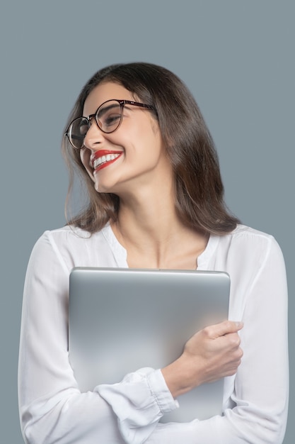 Bonheur, émotion. Enthousiaste jeune femme à lunettes et chemisier tenant un ordinateur portable à son debout sur fond clair