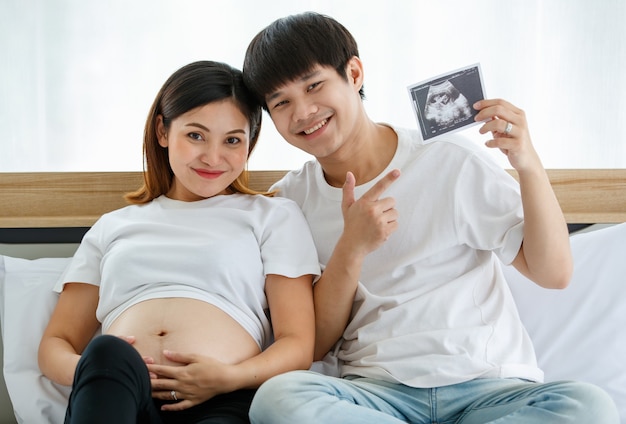 Bonheur jeune couple asiatique vêtu d'une robe décontractée souriant et assis sur un lit ensemble. Un mari heureux met son bras autour de sa femme enceinte et montre une photo d'échographie d'un fœtus.