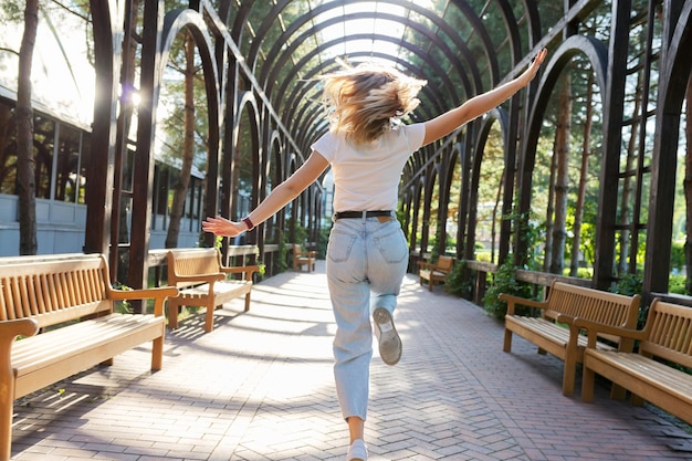 Bonheur, joie, mouvement, liberté, concept de jeunesse. Jeune femme en cours d'exécution à bras ouverts, vue arrière, cheveux au vent au soleil, journée d'été dans le parc, allée voûtée en bois