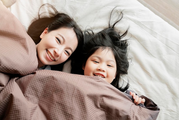 Bonheur moment de famille asiatique maman et fille jouent et se couchent avec une couverture sur fond intérieur de maison de lit blanc