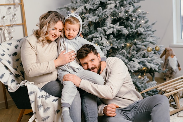 Bonheur de Noël en famille Portrait de papa, maman enceinte et petit fils assis dans un fauteuil à la maison près de l'arbre de Noël câlin sourire européen jeune adulte matin de vacances en famille