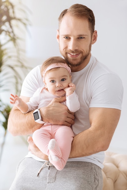 Le bonheur de la paternité. Joyeuse petite fille concentrée mignonne se trouvant dans les mains du jeune père et regardant ailleurs tout en exprimant la positivité