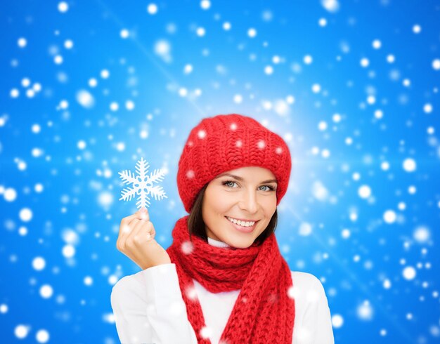 bonheur, vacances d'hiver, noël et concept de personnes - jeune femme souriante au chapeau rouge, écharpe et mitaines tenant un flocon de neige sur fond de lumières bleues