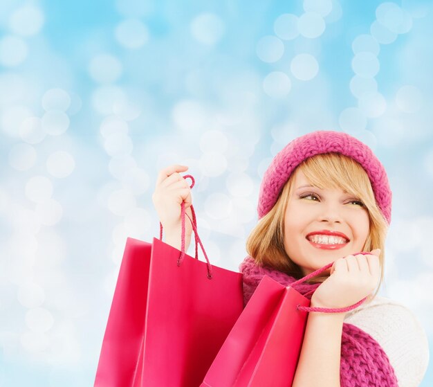 bonheur, vacances d'hiver, noël et concept de personnes - jeune femme souriante en bonnet et écharpe avec des sacs à provisions roses sur fond de lumières bleues