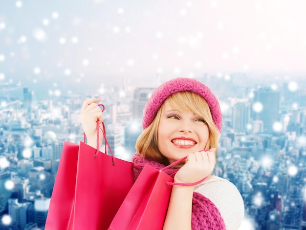 bonheur, vacances d'hiver, noël et concept de personnes - jeune femme souriante en bonnet et écharpe avec des sacs à provisions roses sur fond de ville enneigée