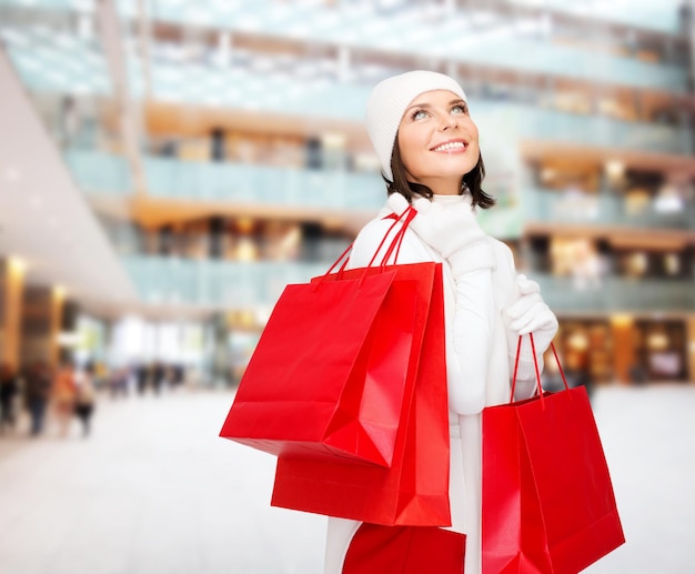 bonheur, vacances d'hiver, noël et concept de personnes - jeune femme souriante en chapeau blanc et mitaines avec des sacs à provisions rouges sur fond de centre commercial