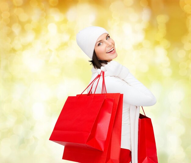 bonheur, vacances d'hiver, noël et concept de personnes - jeune femme souriante en chapeau blanc et mitaines avec des sacs à provisions rouges sur fond de lumières jaunes