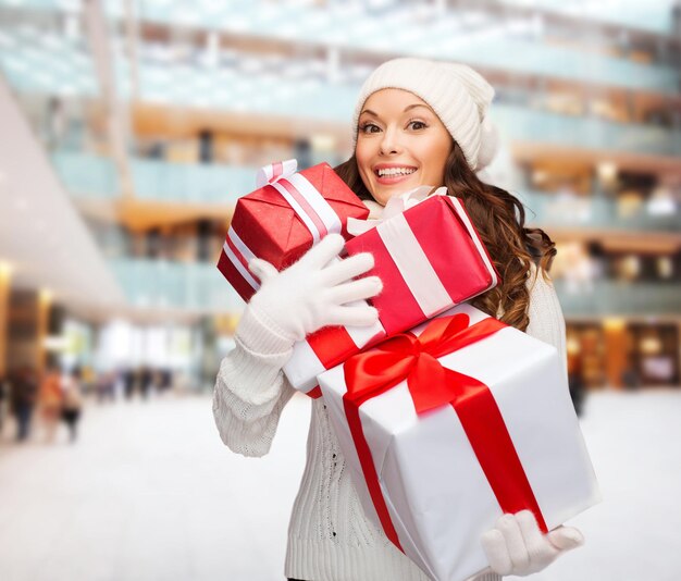 bonheur, vacances d'hiver, noël et concept de personnes - jeune femme souriante en chapeau de santa helper avec des cadeaux sur fond de centre commercial