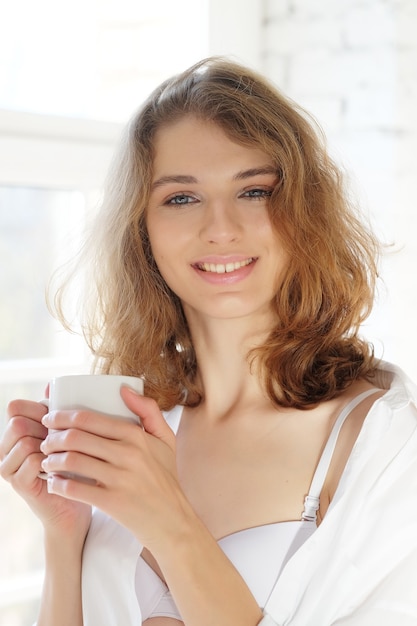 Bonjour ! Belle femme en lingerie blanche buvant du café le matin assise près de la fenêtre.