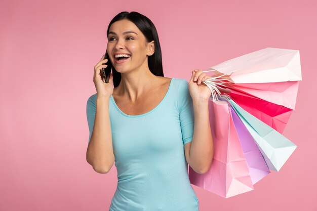 Bonjour, grosses ventes. Portrait de femme discutant au smartphone et tenant des sacs en papier, des achats et des prix agréables. Studio intérieur tourné isolé sur fond rose