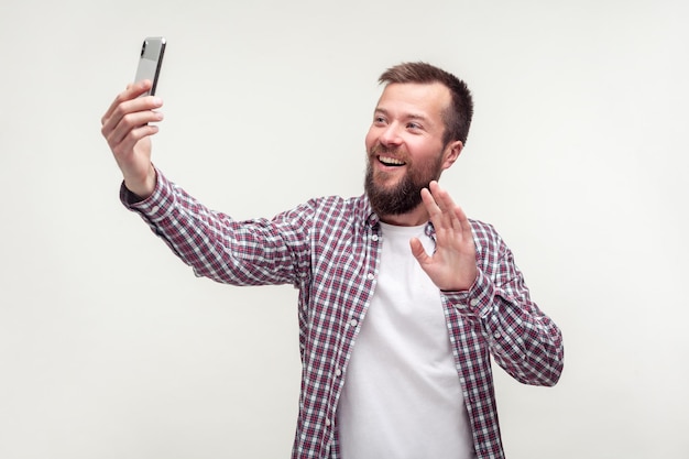 Bonjour Portrait d'un bel homme barbu positif en chemise à carreaux décontractée parlant par appel vidéo à l'aide d'un smartphone et agitant la main en faisant un geste salut studio de communication mobile tourné sur fond blanc