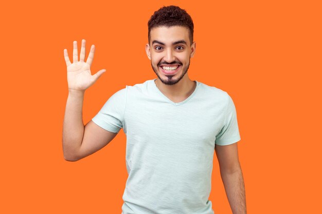 Bonjour. Portrait d'un bel homme brune sympathique et heureux avec une barbe en t-shirt blanc décontracté montrant un geste de salut avec la main agitant et souriant sincèrement. studio d'intérieur tourné isolé sur fond orange