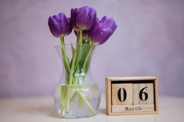 Bonjour printemps Calendrier cubique en bois avec date du 6 mars entouré d'un bouquet de tulipes violettes avec des feuilles vertes dans un vase en verre