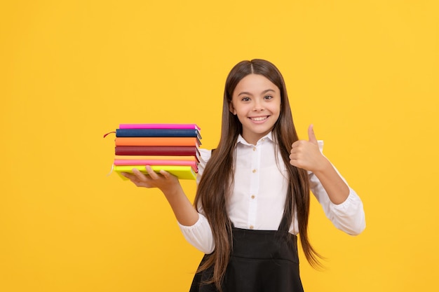 Bonne adolescente en uniforme scolaire tenir la pile de livres montrer le geste du pouce vers le haut, l'excellence.