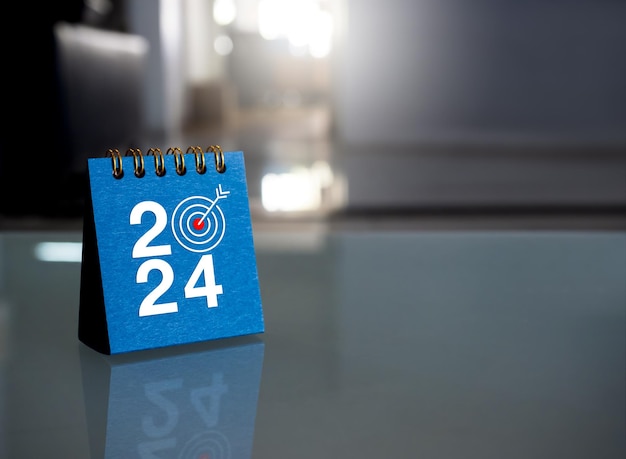 Bonne année 2024 fond de bannière 2024 numéros année avec icône cible sur la couverture bleue du petit calendrier de bureau debout sur une table en verre au bureau avec espace de copie Objectifs commerciaux et concepts de réussite