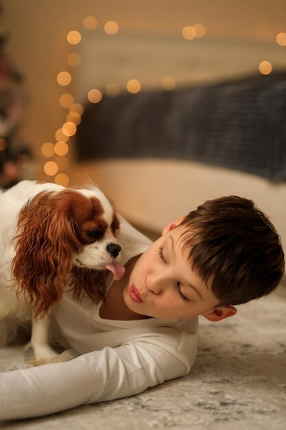 Bonne année un garçon en pyjama léger fait maison embrasse son animal de compagnie cavalier king charles spaniel à la maison dans la chambre près de l'arbre de Noël