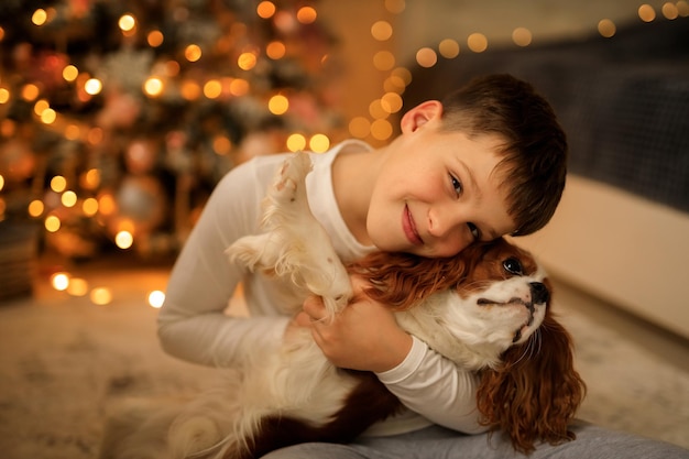 Photo bonne année un garçon en pyjama léger fait maison étreint son animal de compagnie cavalier roi charles spaniel à la maison dans la chambre près de l'arbre de noël