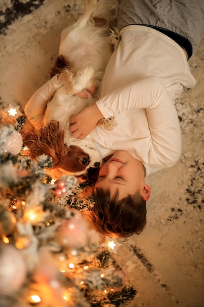 Bonne année un garçon en pyjama léger fait maison étreint son animal de compagnie cavalier roi charles spaniel à la maison dans la chambre près de l'arbre de Noël