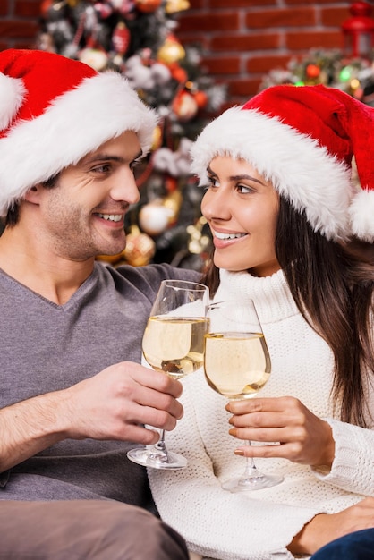 Bonne année! Heureux jeune couple d'amoureux en chapeaux de Père Noël se liant les uns aux autres et tenant des verres à vin avec un arbre de Noël en arrière-plan